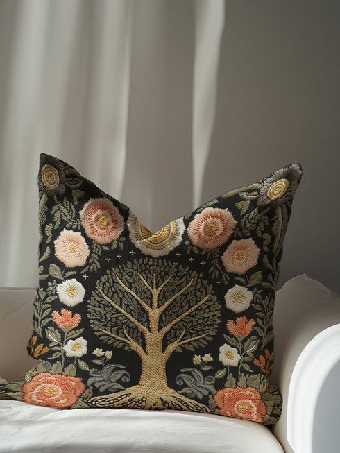 A square charcoal black decorative pillow cover made from a linen-feel fabric. The design features a large, central Tree of Life in antique gold and sage green, with intricate roots at the base. The tree is surrounded by a symmetrical pattern of stylized folk art flowers in muted blush pink, terracotta, and off-white. The print has a faux embroidered, tapestry-style texture. In each of the four corners is a circular, sun-like medallion, completing the ornate, botanical noir design.